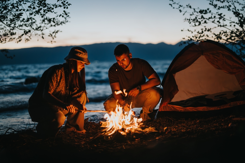 Two people by campfire near a lake and a tent