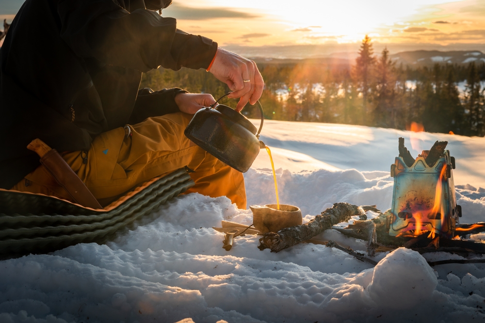 Pouring a cup of coffee from a kettle brewed on a camp fire in winter