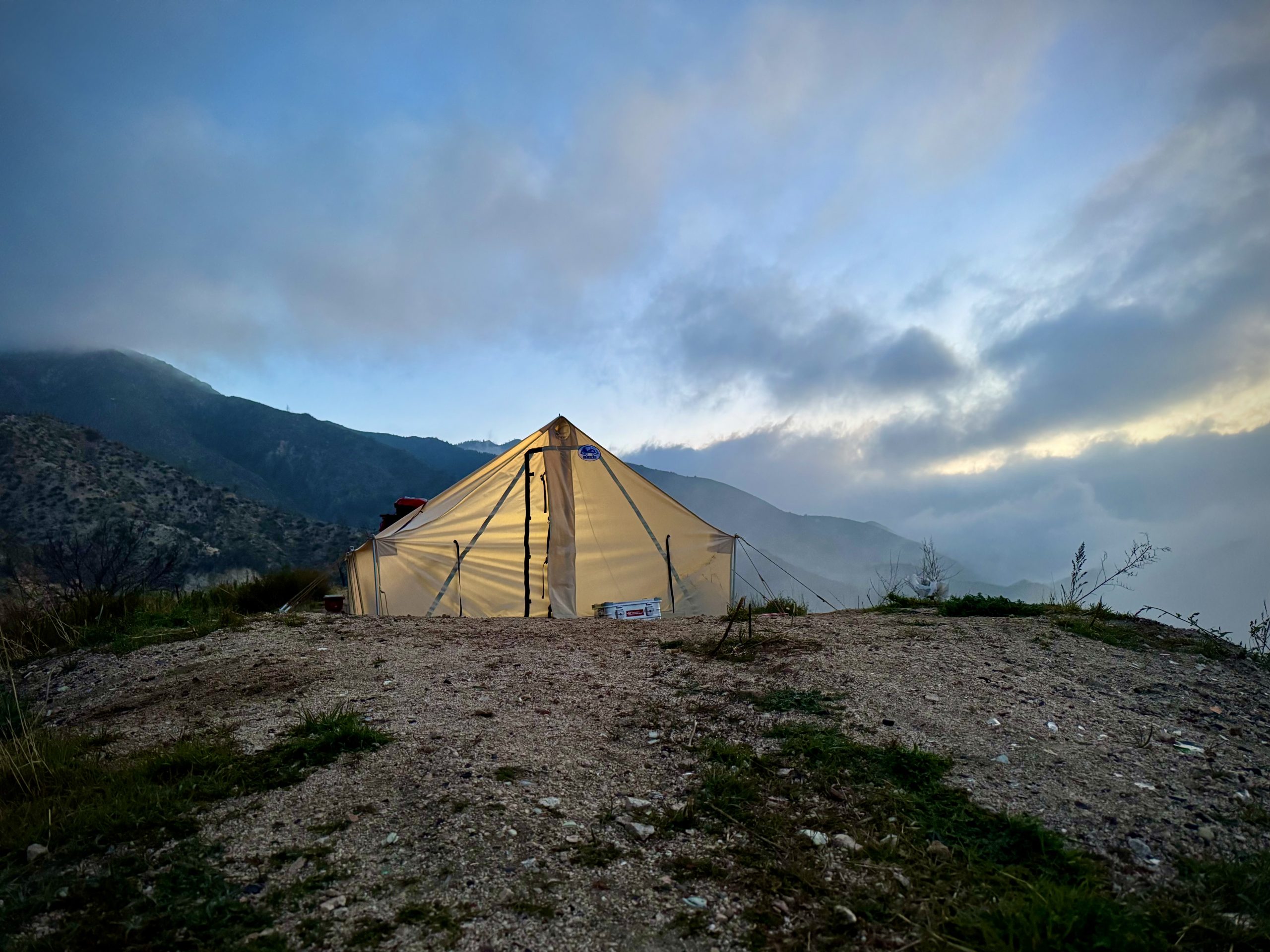 Sunforger canvas tent glowing at dusk on a gravel ridge with layered mountain silhouettes behind