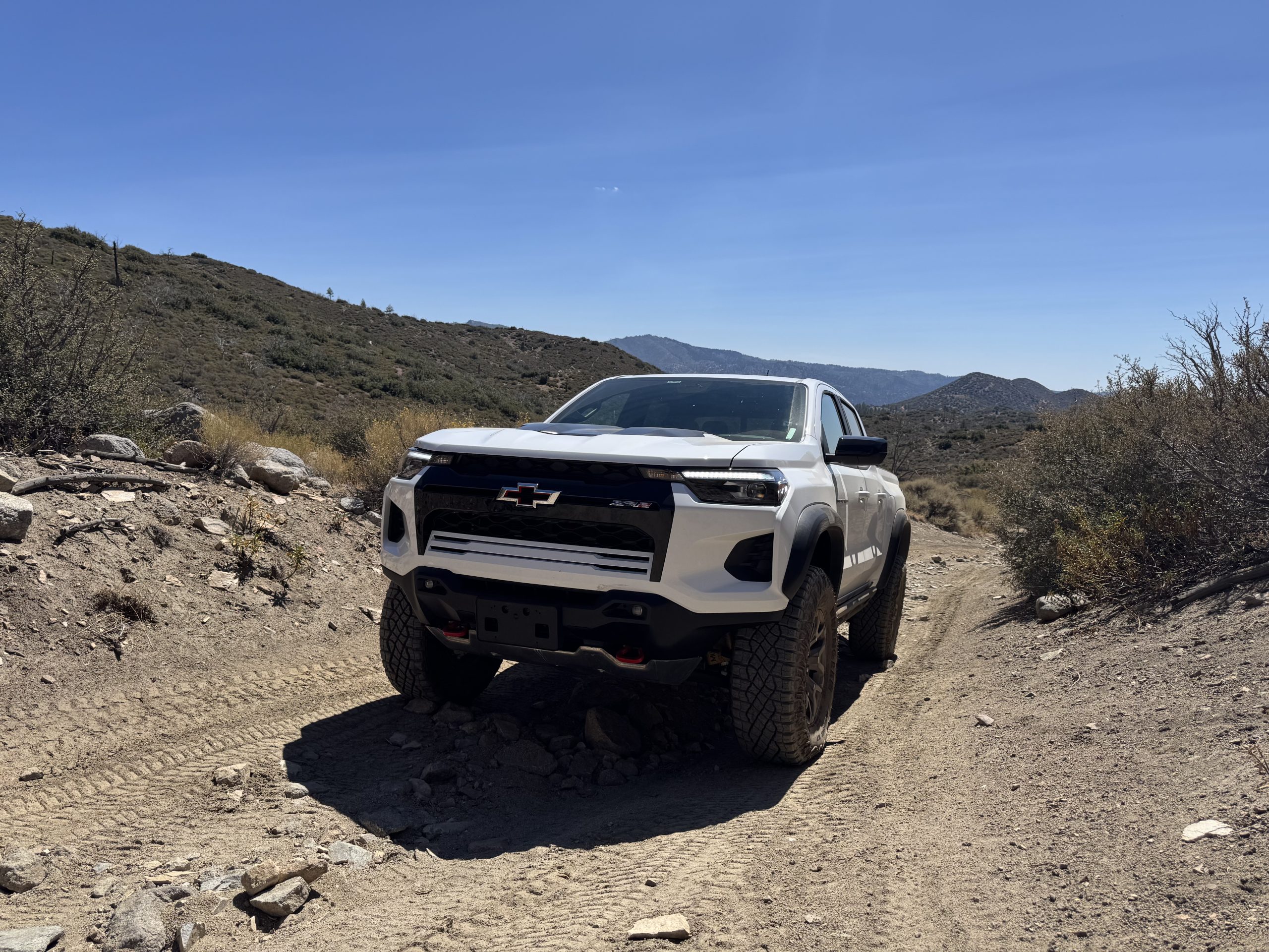 White Chevy Colorado ZR2 climbing a rocky dirt trail on a chaparral-covered ridge with mountains in the distance