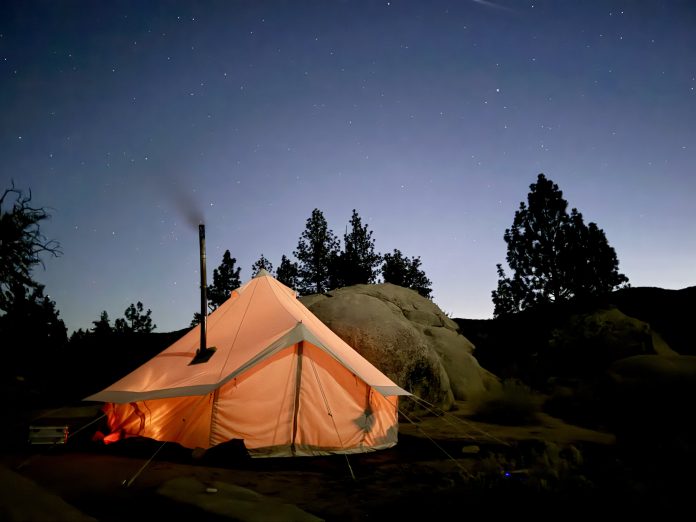 Illuminated orange canvas bell tent with a hot tent stove pipe glowing under a starry night sky near boulders