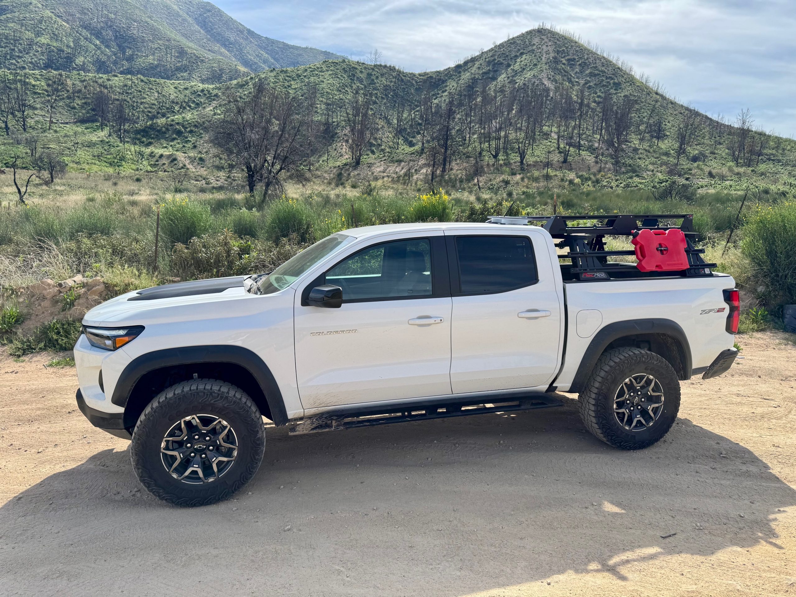 White Chevy Colorado ZR2 with roof rack and red fuel canister parked beside green hills after a wildfire burn