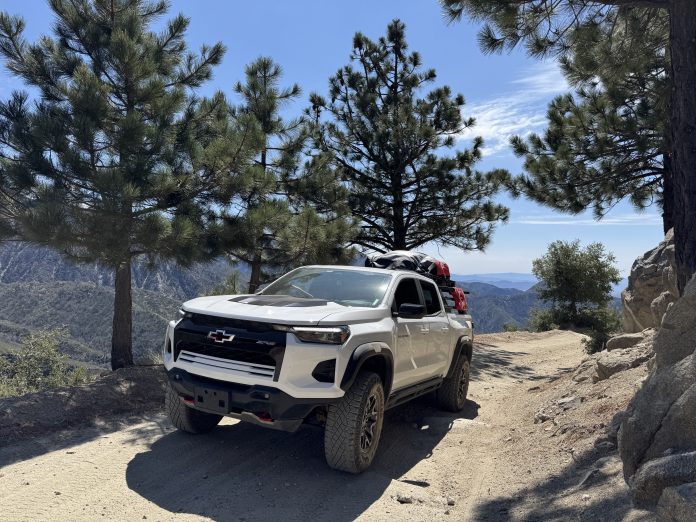 White Chevy Colorado ZR2 with roof rack and gear parked on a pine-lined mountain overlook trail