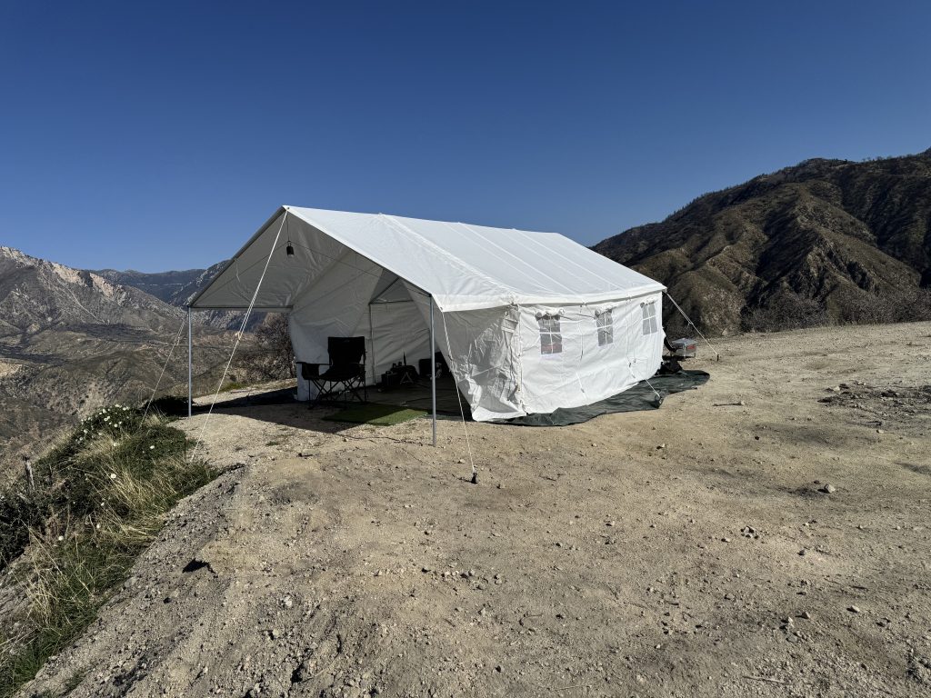 White canvas wall tent with window panels set up on a dirt overlook above chaparral canyons under clear sky