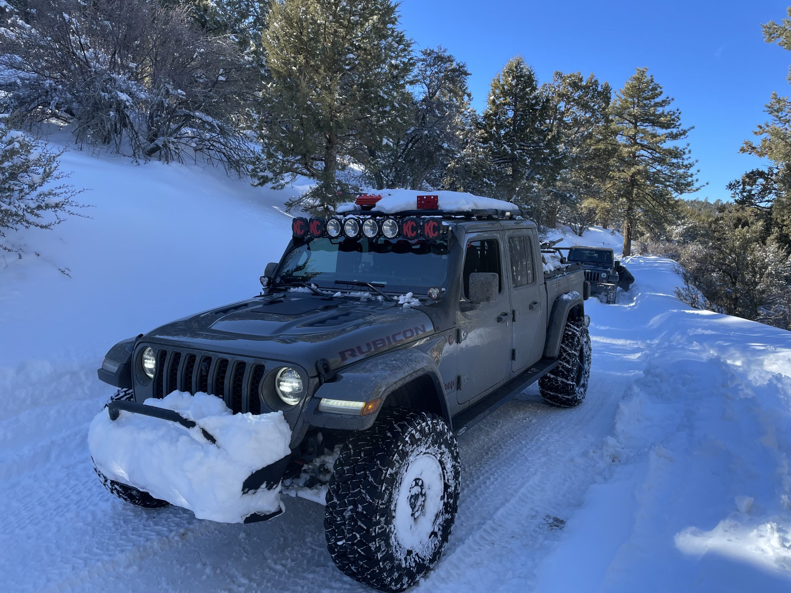 Jeep Gladiator Rubicon with snow-covered hood and grille driving a snowy mountain trail past another Jeep