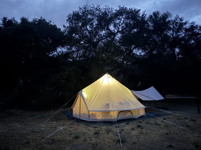 Stout 5M Pro bell tent glowing from interior light at dusk beneath oak trees at a campsite.
