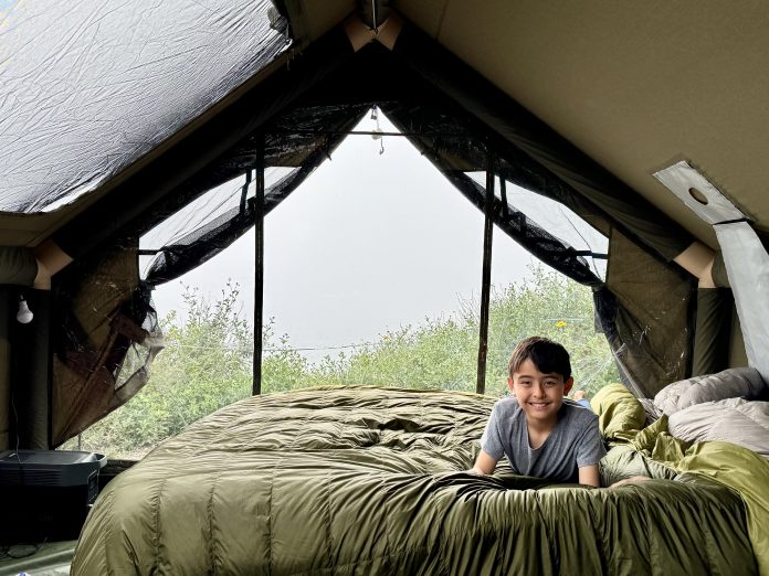 Boy smiling on an Aeronaut Hoverquilt inside a rooftop tent with a mesh window view of greenery outside.