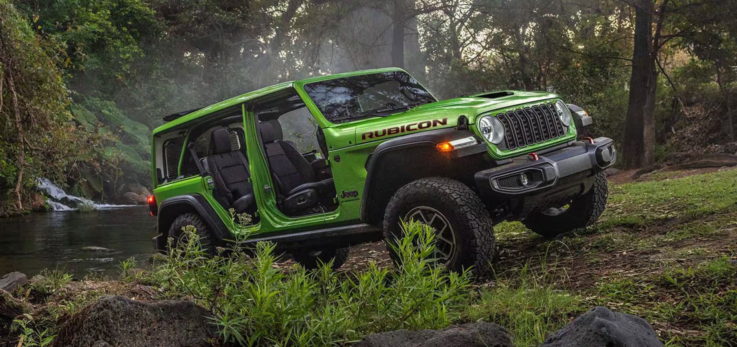 Green Jeep Wrangler Rubicon two-door with doors off, parked beside a forest stream and small waterfall — manufacturer image