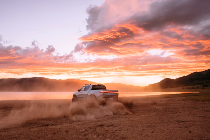 White Rivian R1T truck kicking up dust beside a lake under a vivid orange sunset sky — manufacturer image