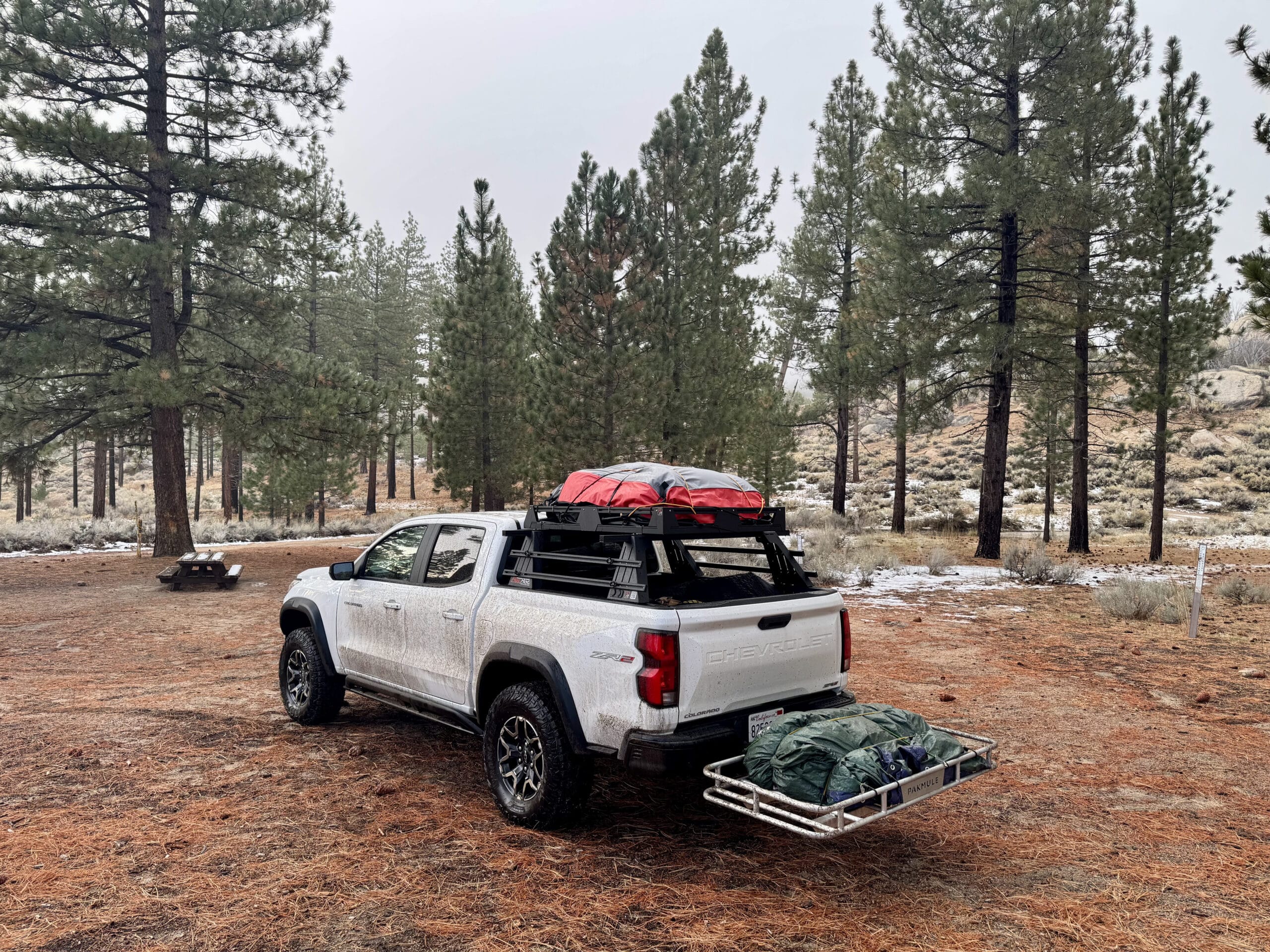 White Chevy Colorado ZR2 with rooftop tent and Pakmule cargo rack loaded with gear at a snowy pine campsite