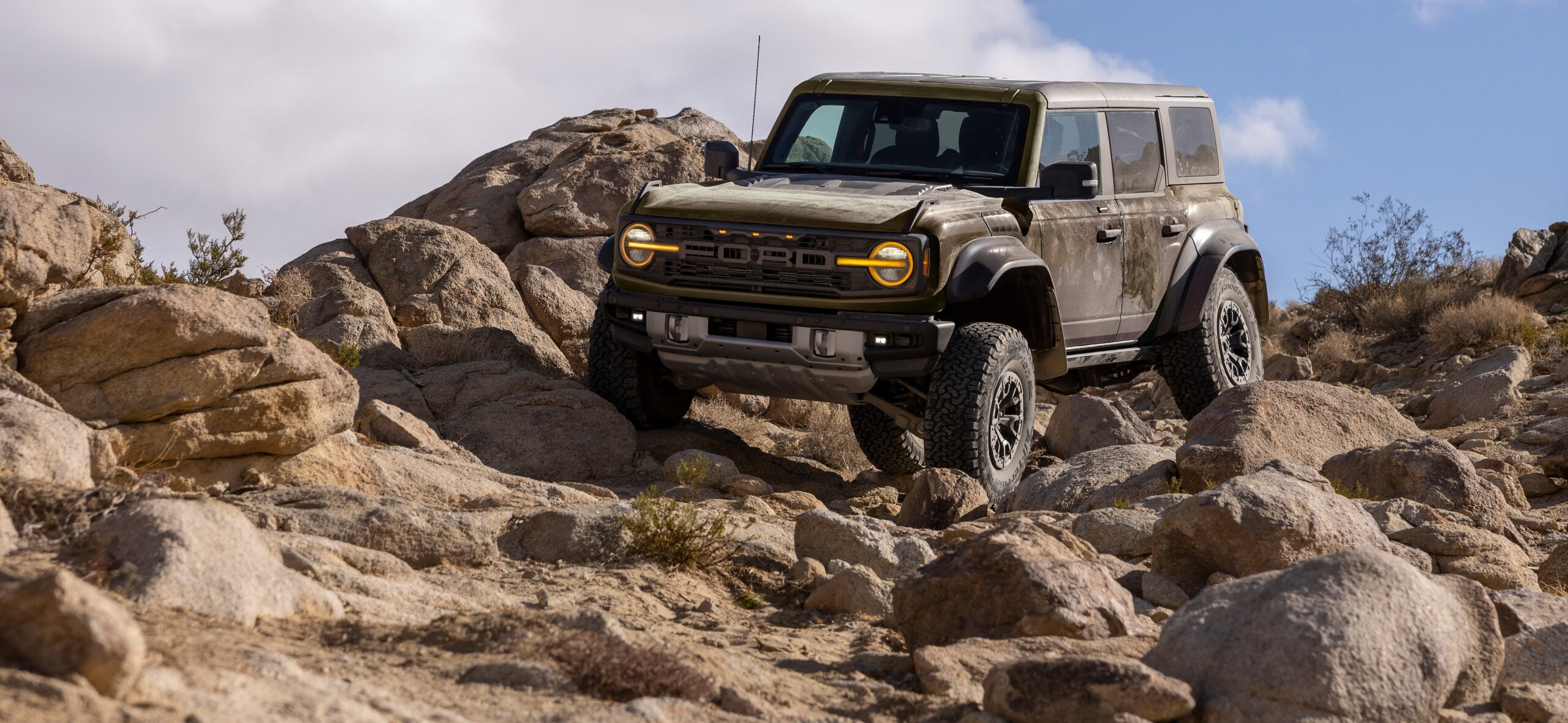 Shelter Green Ford Bronco Raptor with amber grille lighting climbing a boulder-strewn rocky hillside trail