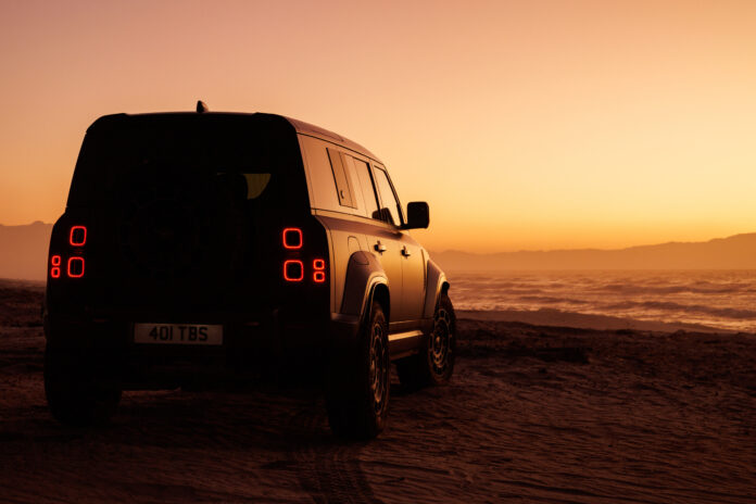 Silhouetted Land Rover Defender Octa parked on a beach at sunset with its taillights glowing red