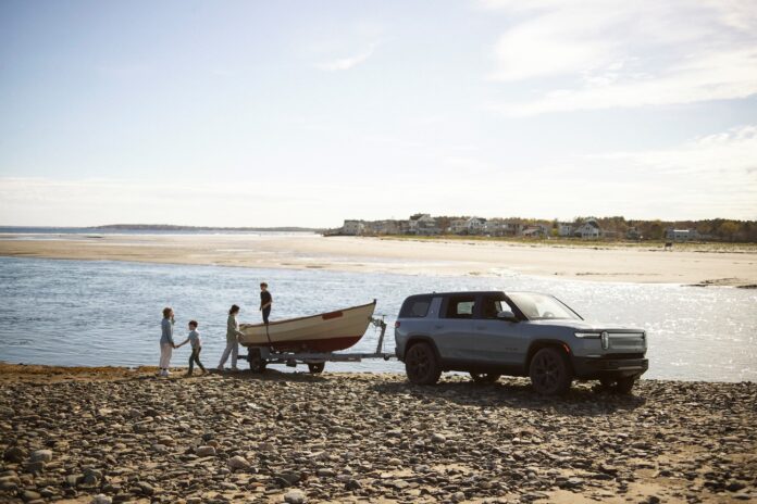 Family launching a wooden rowboat from a trailer behind a gray Rivian R1S parked on a tidal beach