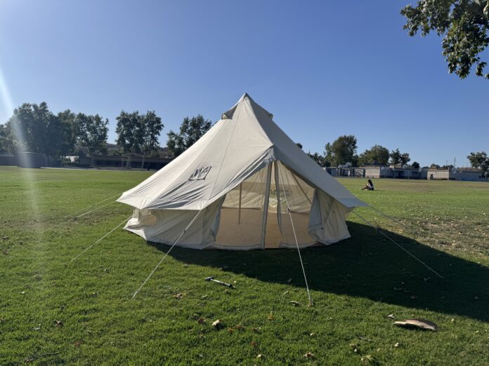 Cream canvas bell tent fully pitched on a grassy field with guy lines staked out and trees in the background