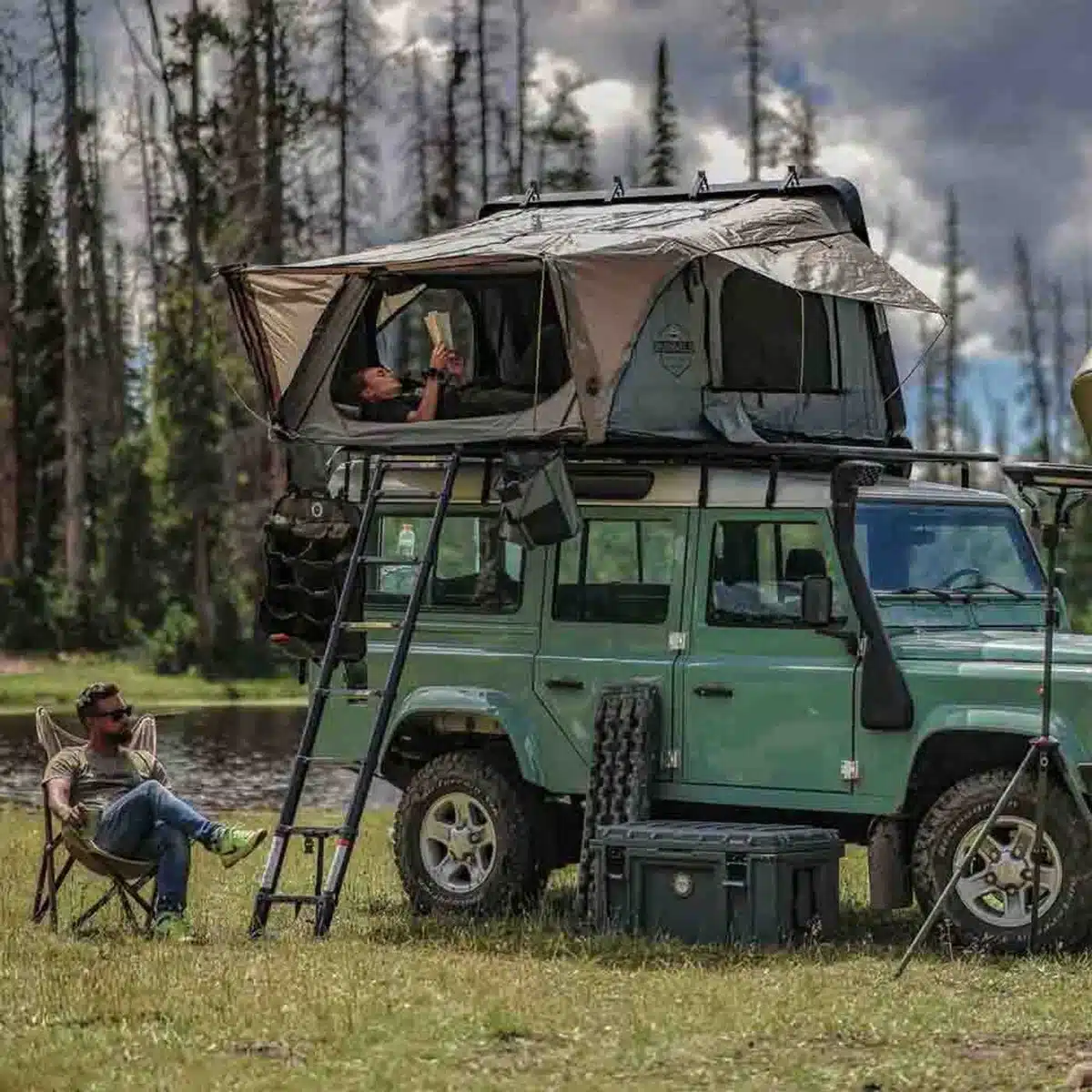 Olive Land Rover Defender with an OVS Bushveld II hard-shell rooftop tent open and a man reading inside