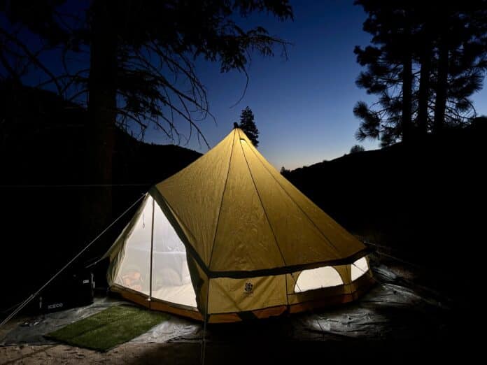 Yellow canvas bell tent glowing from within at dusk among silhouetted pine trees on a hillside