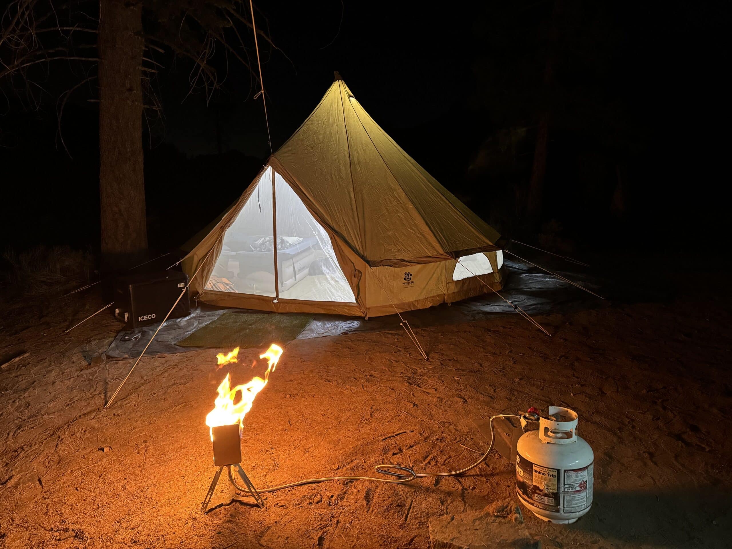Bell tent at night with mesh door open, a Tripod Torch fire burning out front beside a propane bottle