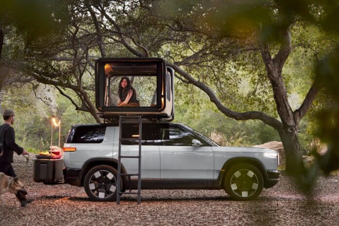 Rivian R2 with pop-up rooftop camper open and a woman inside, campsite cooking gear nearby in the woods