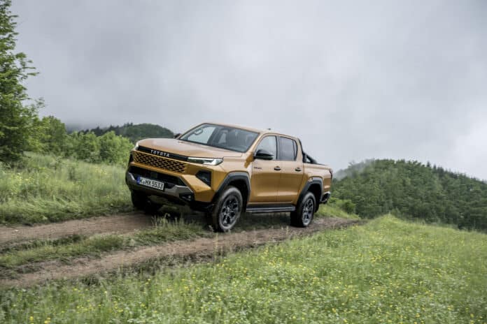 Yellow Toyota Hilux Invincible hybrid pickup climbing a grassy off-road trail under a cloudy sky