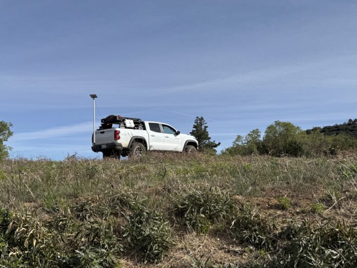 White overland truck loaded with rooftop gear parked on a grassy hilltop under a clear blue sky
