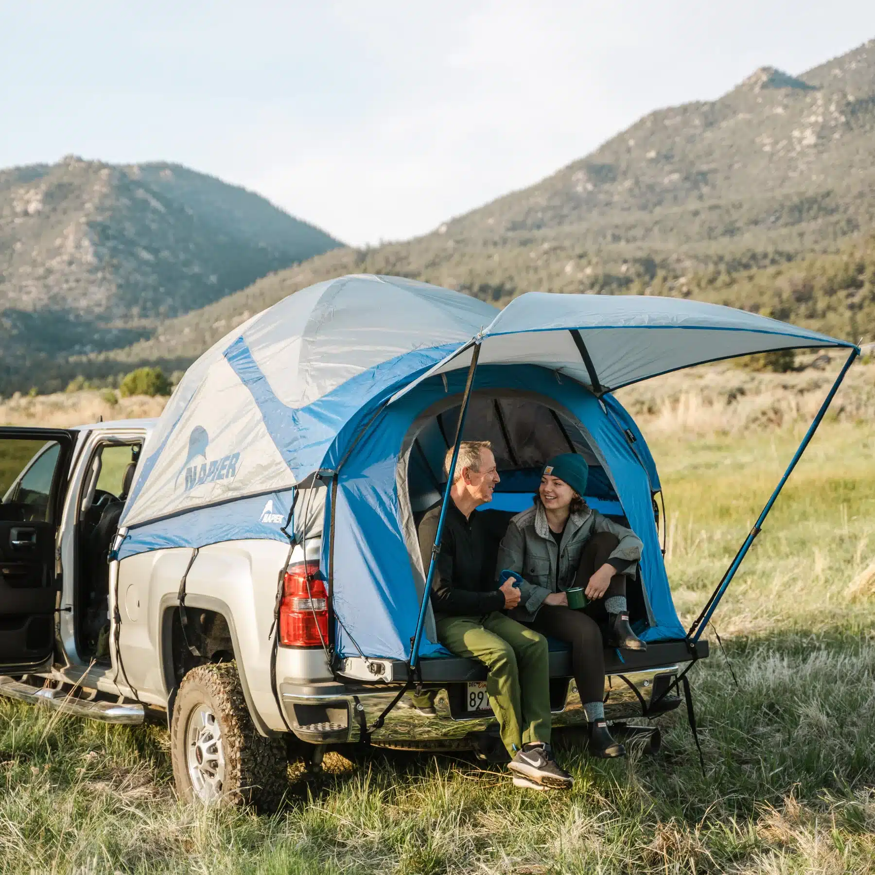 Two people sitting in the tailgate opening of a Napier Sportz truck tent, laughing with mountains behind — manufacturer image