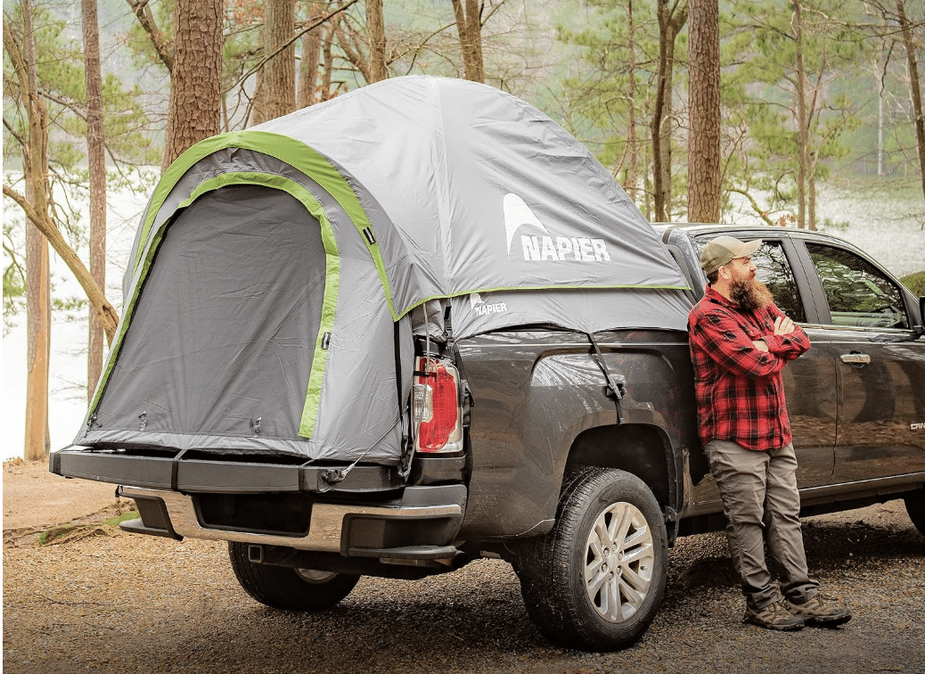 Man standing beside a Napier Backroadz truck bed tent with green trim on a dark pickup at a lakeside campsite