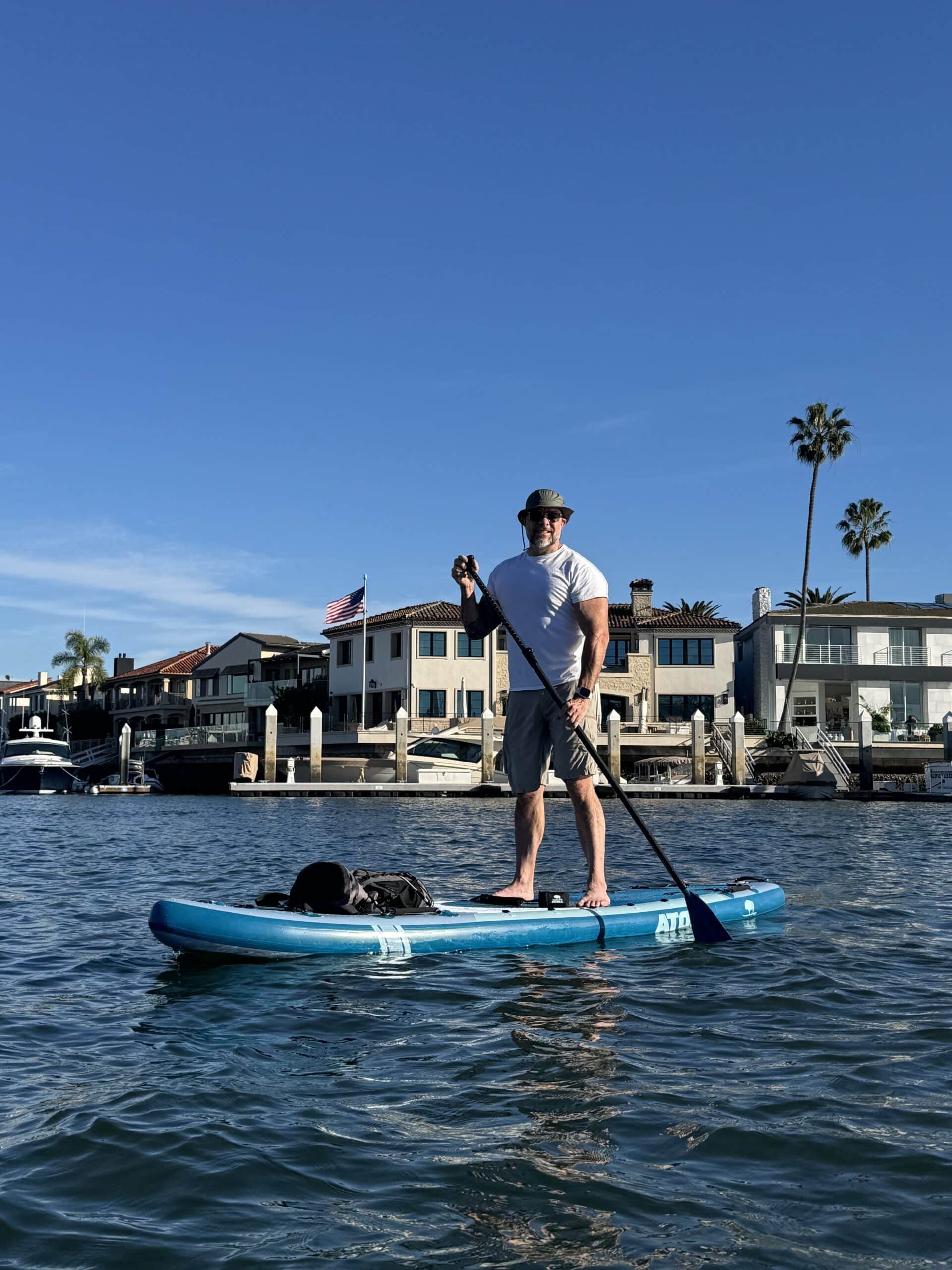 Man in a hat paddling a light blue Atoll Outrider inflatable SUP with dry bag through a marina lined with waterfront homes
