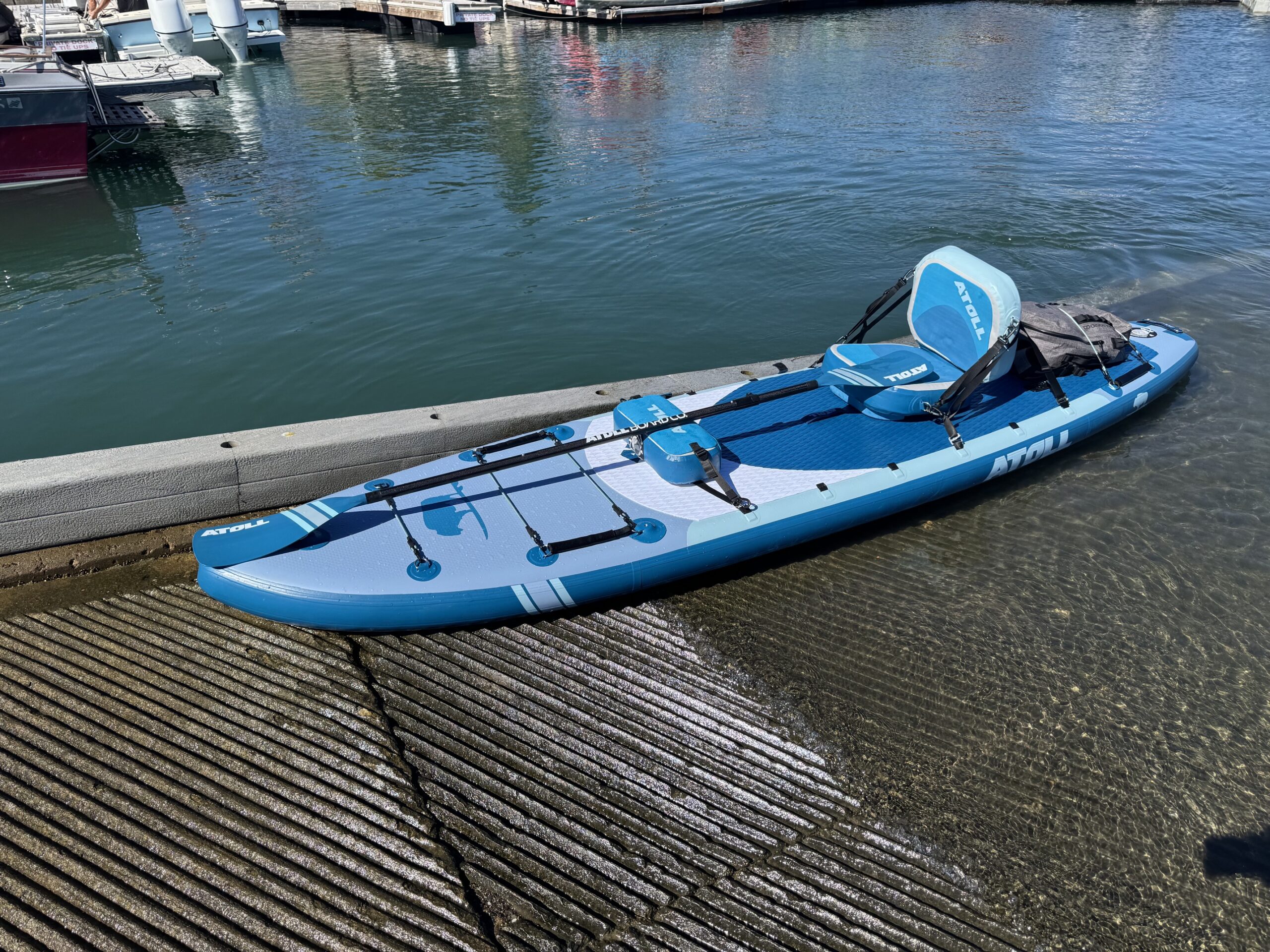 Blue and light blue Atoll Outrider inflatable paddle board with padded seat and cargo straps resting on a boat ramp
