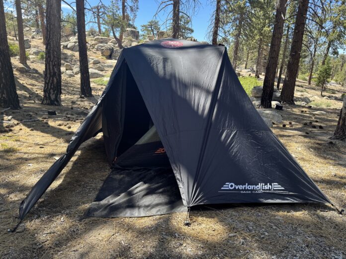 Black Overlandish Base Camp rainfly-style tent pitched among boulders and pine trees in bright sunlight