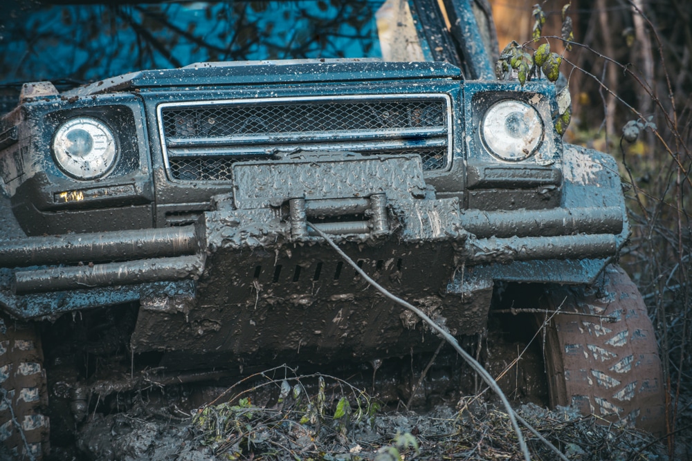 Mud-caked off-road vehicle front end with winch bumper stuck deep in a muddy rut, cable running through the mire