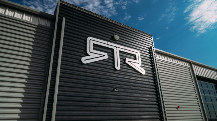 Polished metal RTR Vehicles logo mounted on the dark corrugated siding of the Farmington headquarters