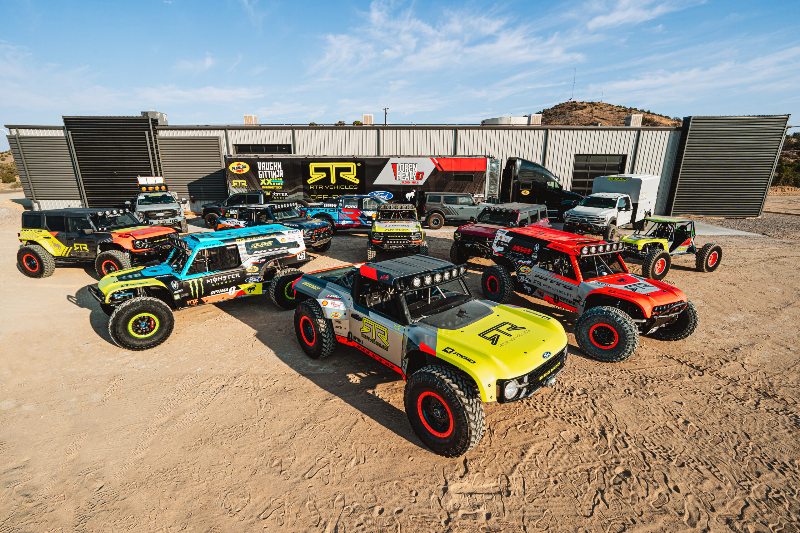 RTR Ultra4 Bronco race trucks, chase trucks, and the team transporter staged on sand outside the Farmington shop