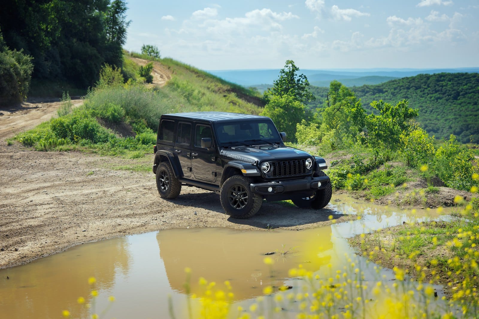 Black 2027 Jeep Wrangler Smoky Mountain on a dirt track above a green valley, standard 33-inch all-terrain tires and 17-inch black wheels in view