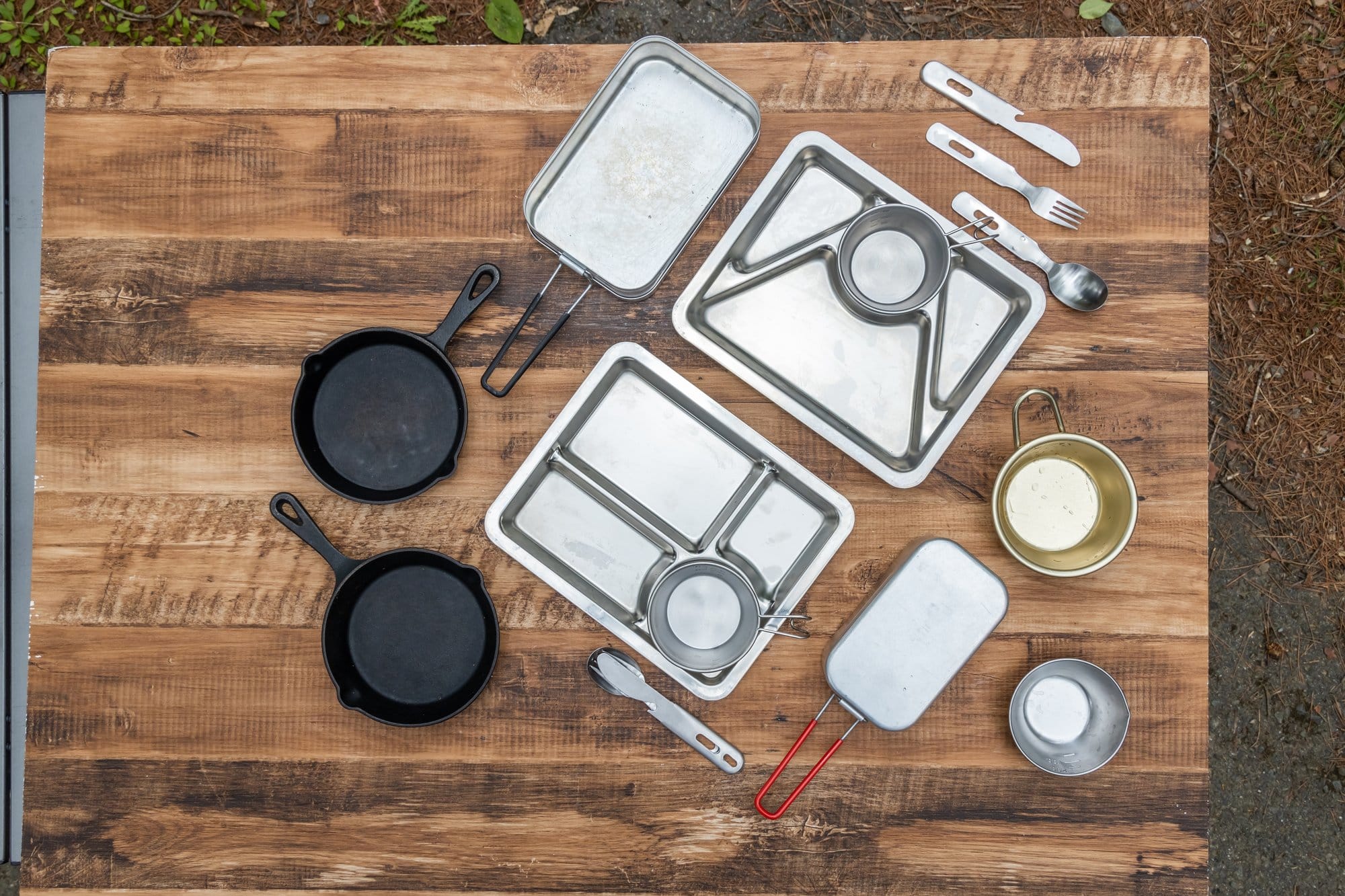 Camp cookware metals compared Cast iron skillets, aluminum mess kits and steel camp cups laid out on a wooden table
