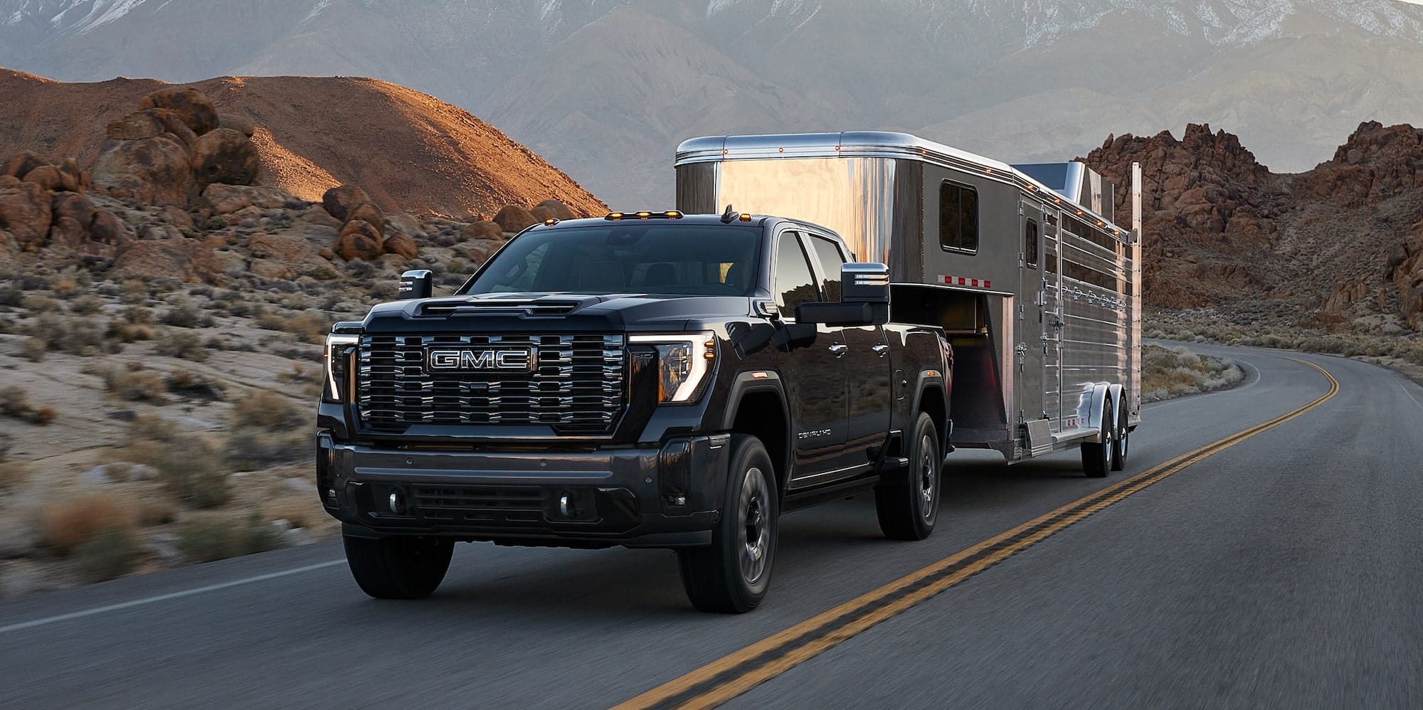 Black 2027 GMC Sierra HD towing a silver horse trailer on a desert highway