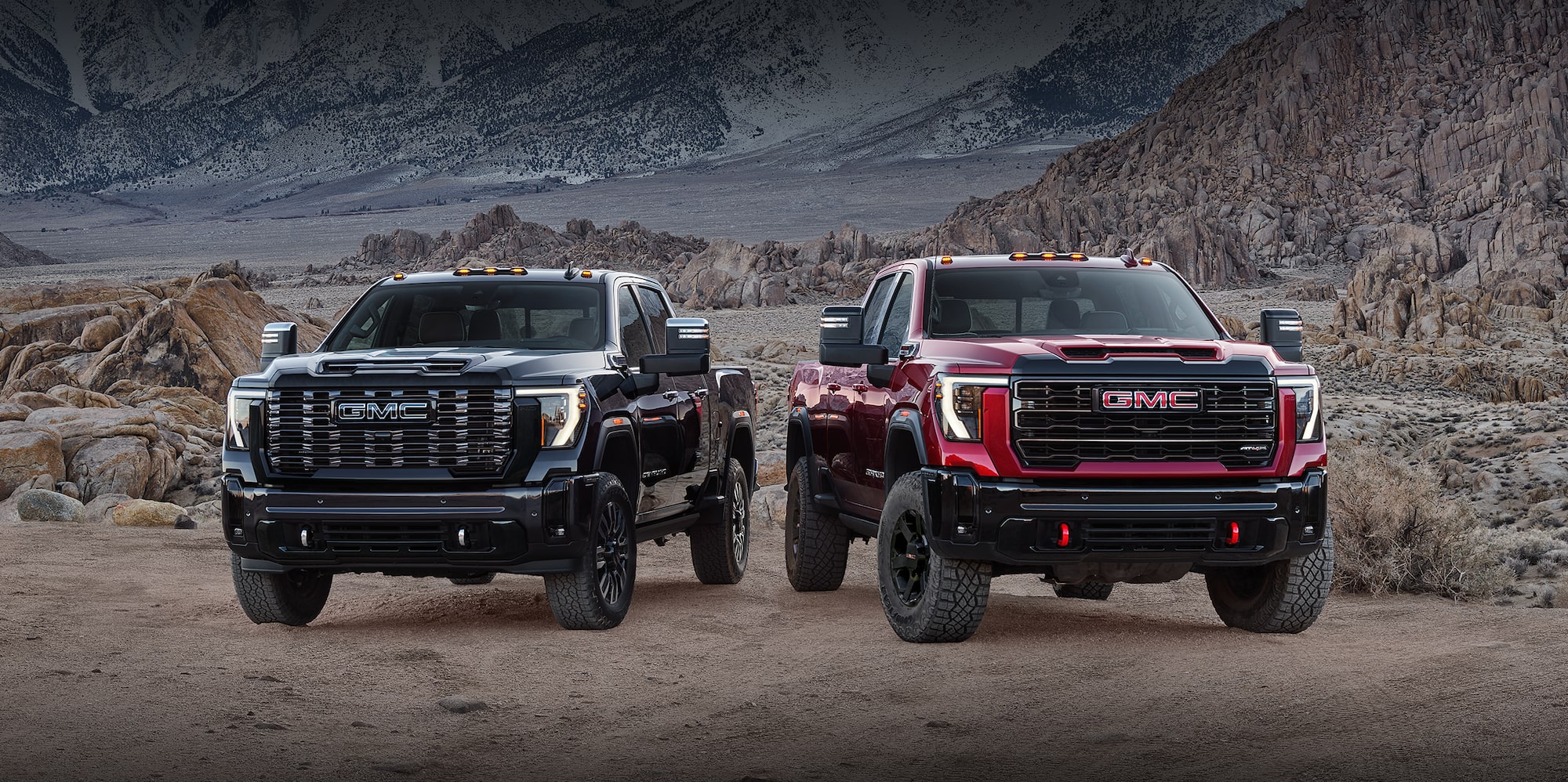 Black and red 2027 GMC Sierra HD trucks with off-road tires and red recovery hooks on rocky terrain
