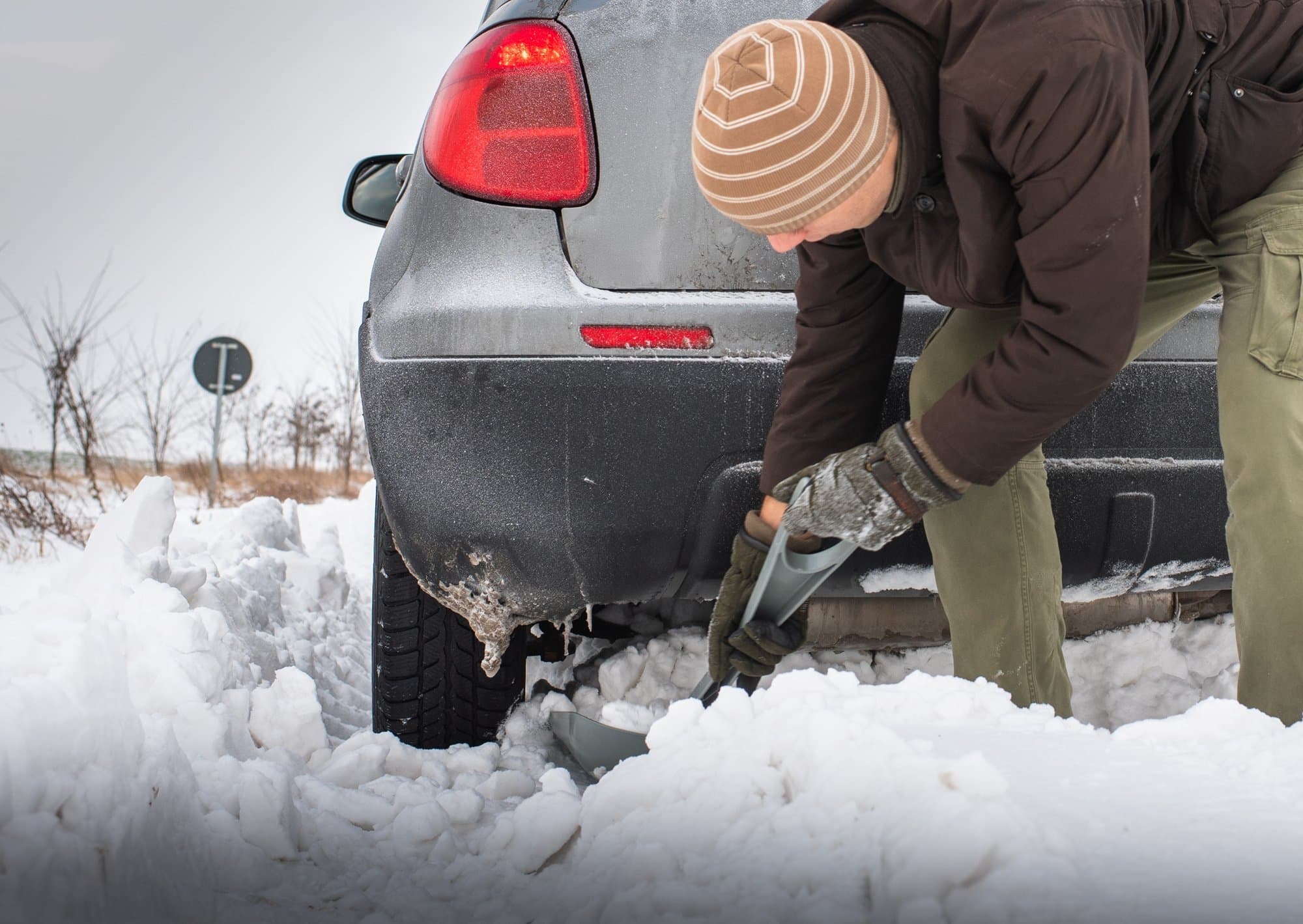 Driver digging snow away from the rear tire of a stuck vehicle with a shovel