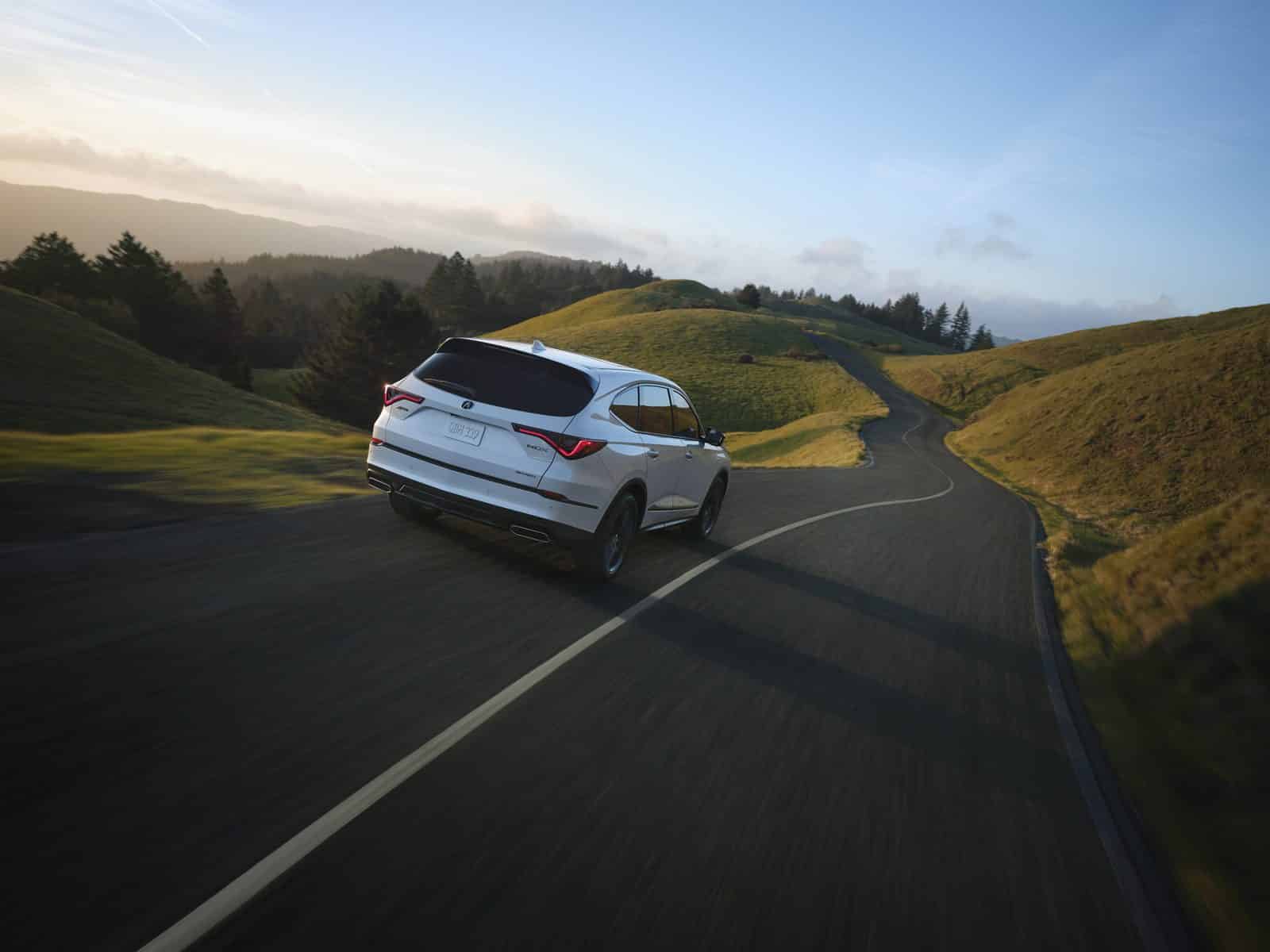 Acura MDX on a winding road White Acura MDX seen from the rear on a winding paved road through green hills