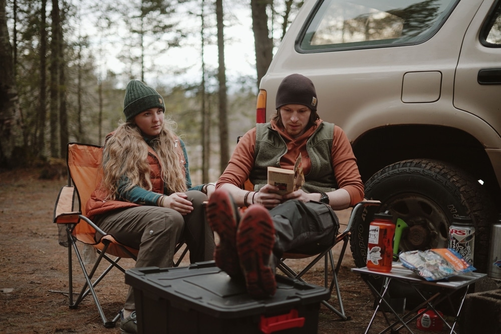 Campers in camp chairs at a cool forest campsite Two campers in beanies and vests sitting in camp chairs beside an SUV at a forest campsite