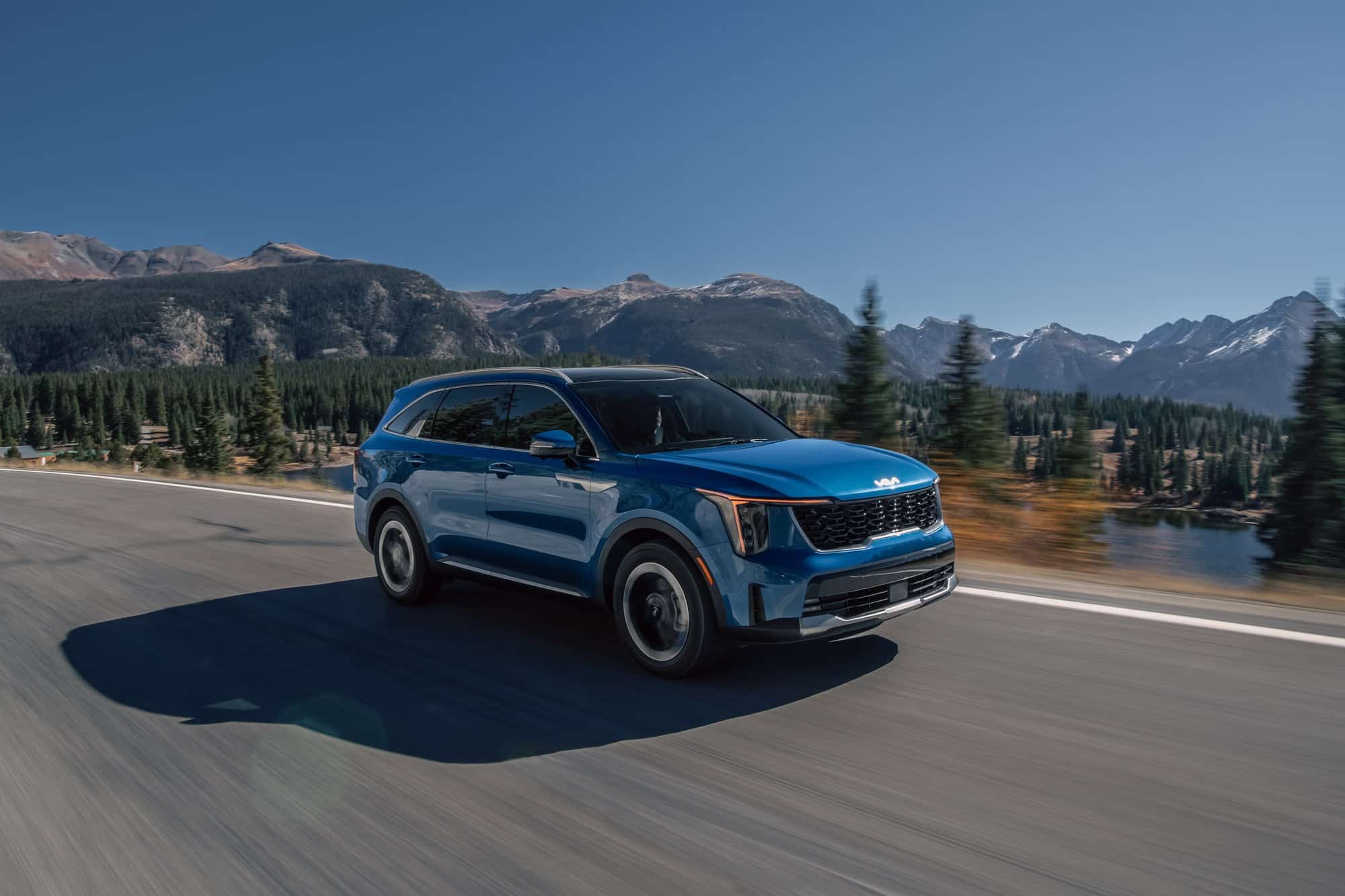 2027 Kia Sorento Hybrid on a mountain road Blue 2027 Kia Sorento Hybrid driving on a mountain highway past pine trees
