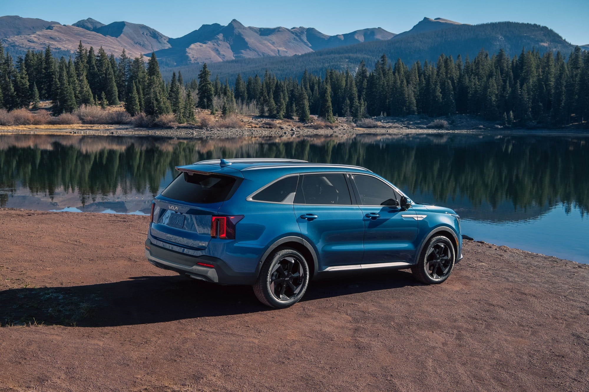 2027 Kia Sorento Hybrid rear view by a lake Rear three-quarter view of a blue 2027 Kia Sorento Hybrid on a dirt shore beside a mountain lake