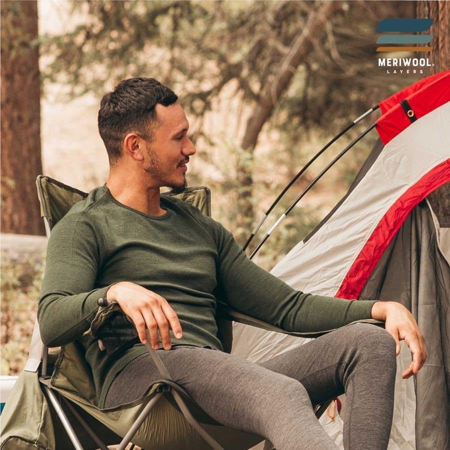 Man in a green MERIWOOL merino wool base layer top and gray bottoms sitting in a camp chair beside a tent