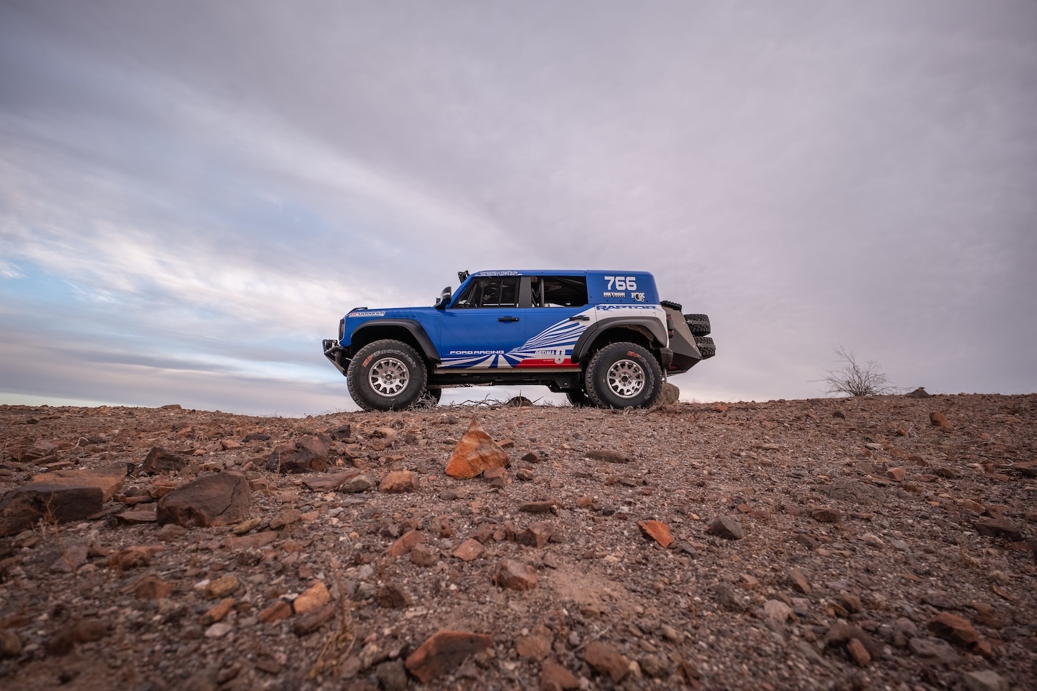 Side view of the blue and white Bronco Raptor Baja race entry on a rocky desert ridge
