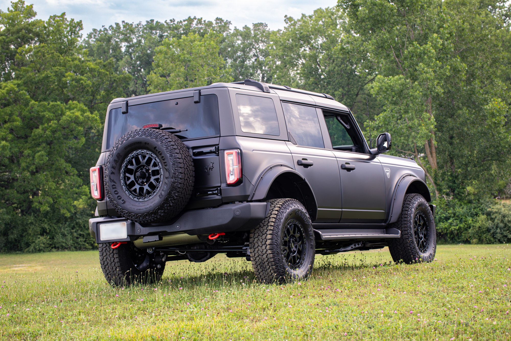 Rear view of a matte black four-door Bronco with ORV Package wheels and tires on grass in front of trees