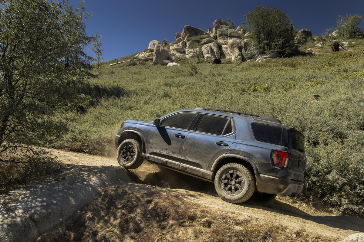 Blue 2027 Honda Passport TrailSport climbing a rocky dirt trail on a brushy hillside