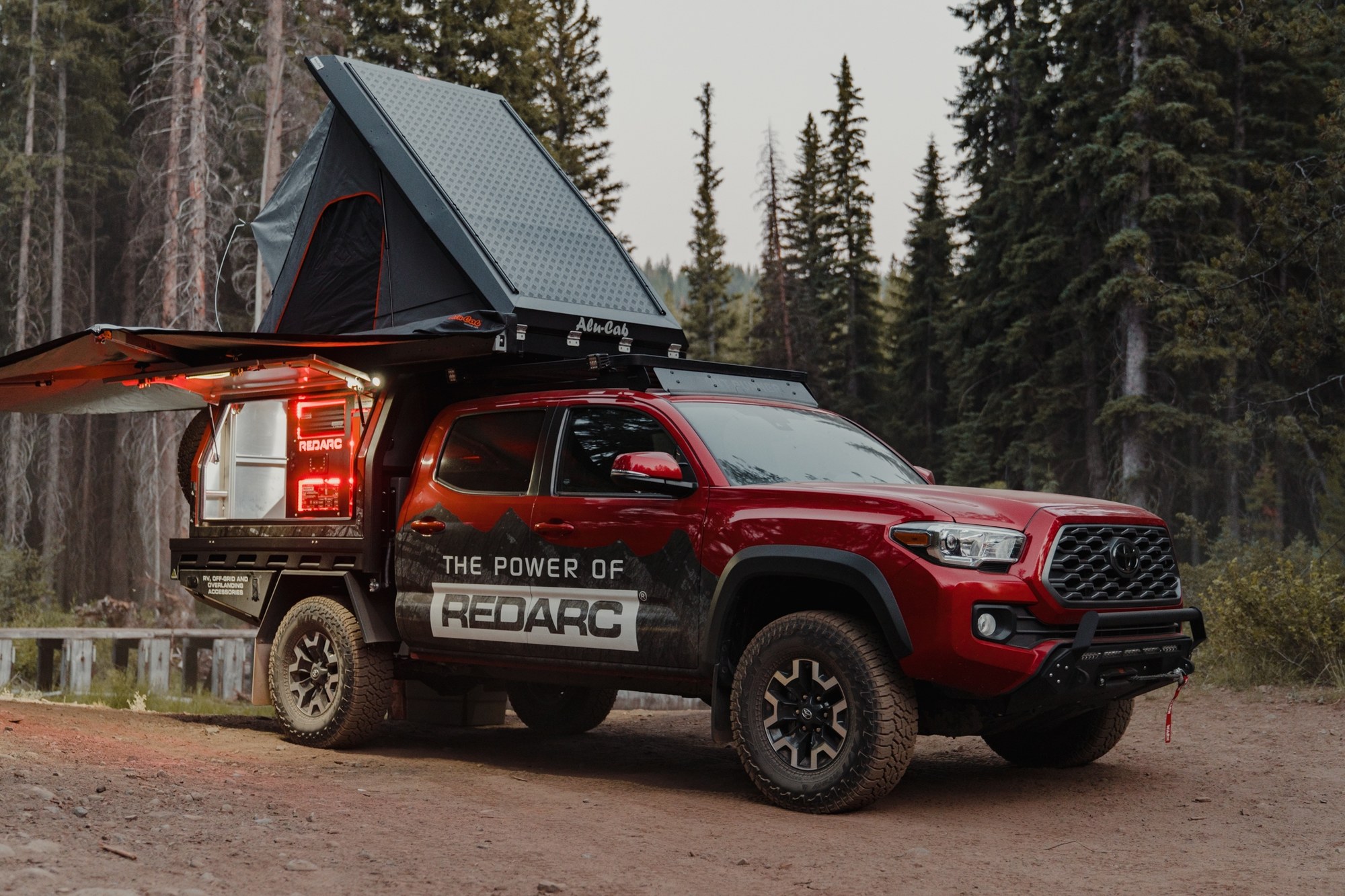 REDARC Tacoma build set up at camp Red Toyota Tacoma with an open rooftop tent and canopy parked at a forest camp