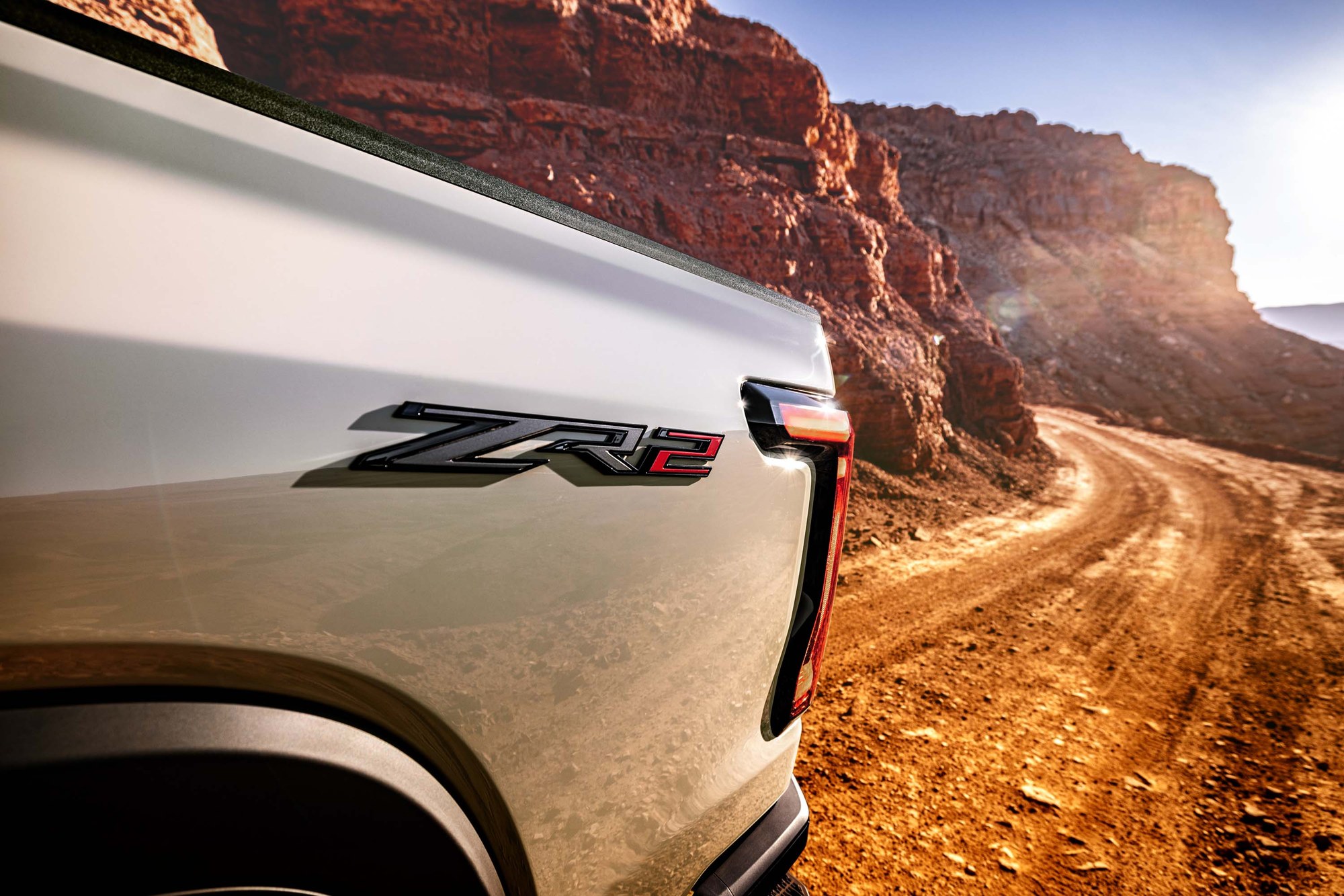 2027 Silverado ZR2 badge on a canyon dirt road ZR2 badge on the bed of a white 2027 Chevrolet Silverado ZR2 beside a dirt road through red rock canyon