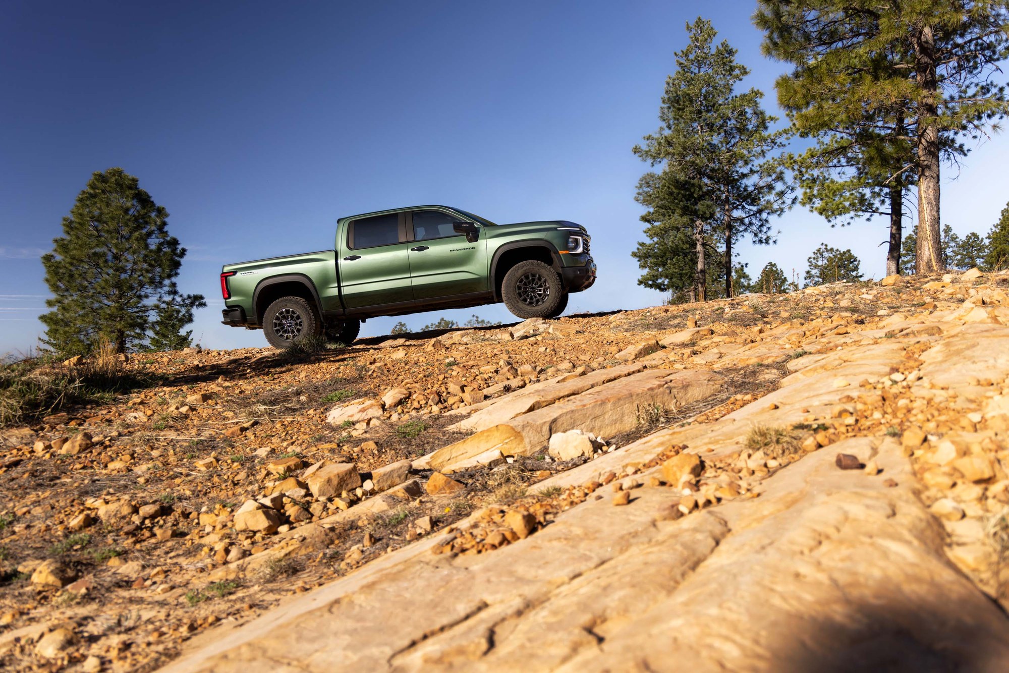 2027 Silverado Trail Boss on a rocky trail Green 2027 Chevrolet Silverado Trail Boss climbing a rocky slickrock trail among pine trees