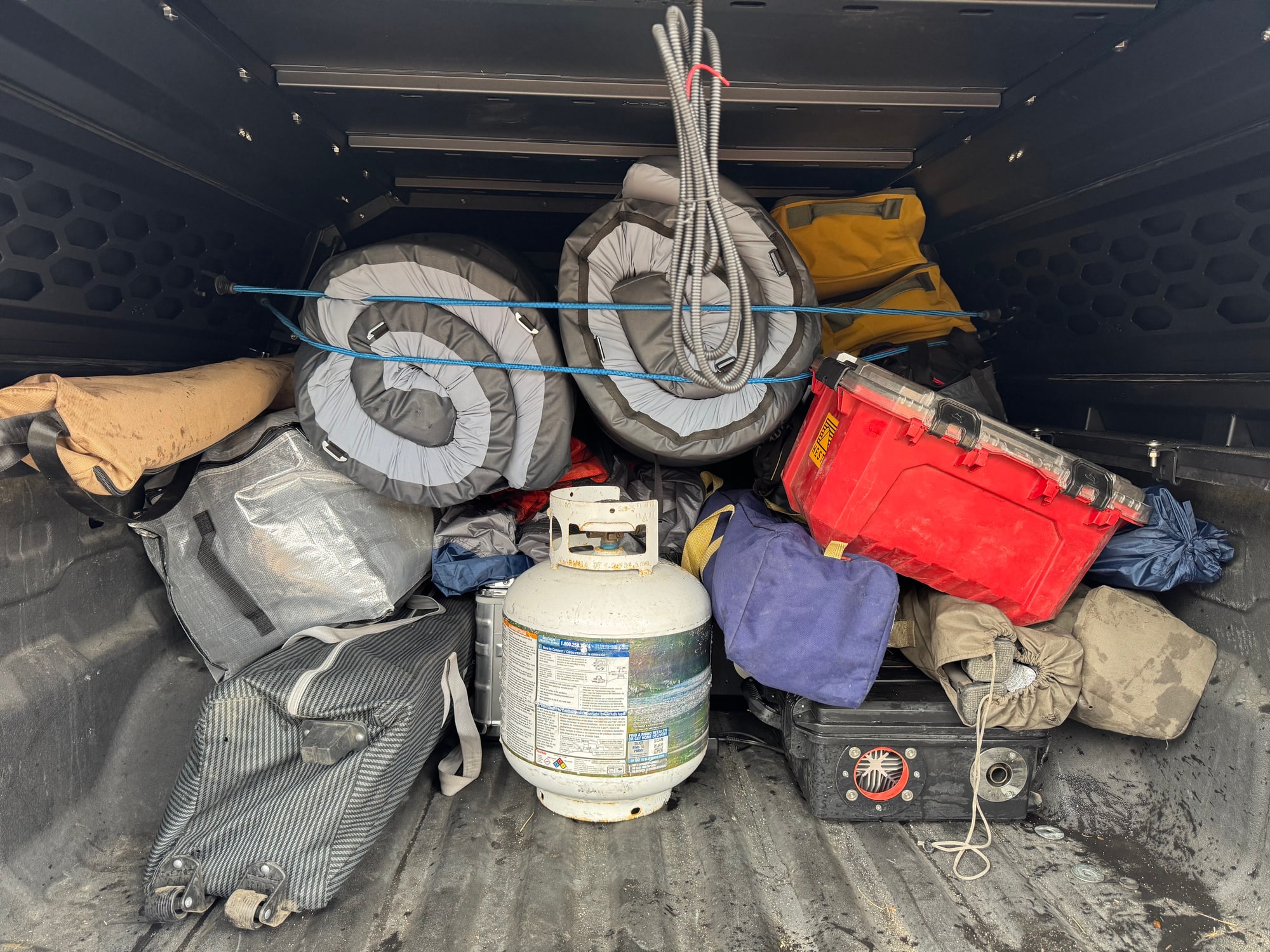 Camping Gear Inside the OPENROAD Truck Cap Truck bed under an OPENROAD cap packed with rolled bedding, bags, a red storage box and a propane cylinder.