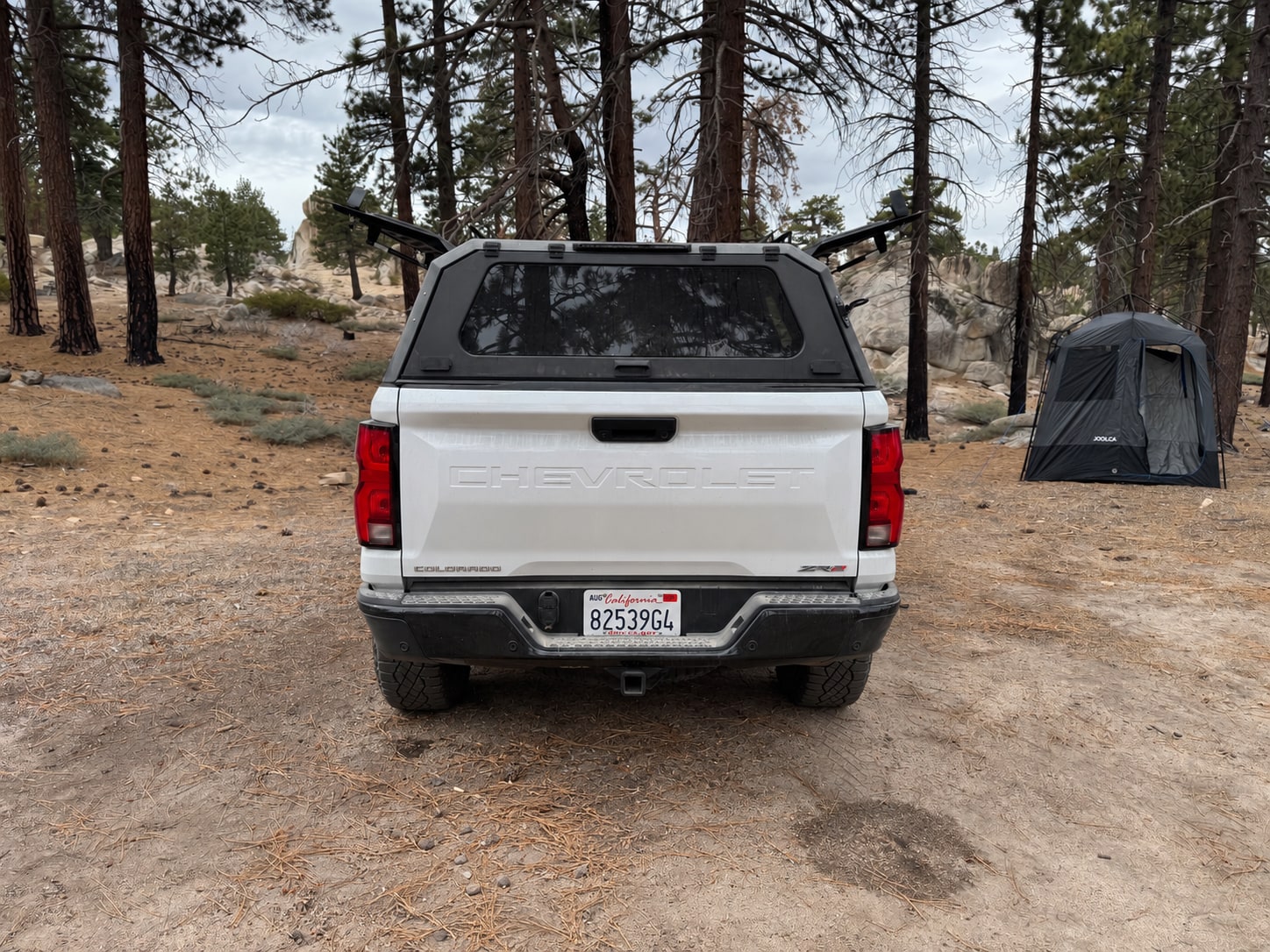OPENROAD Truck Cap Rear View with Side Doors Open Rear view of a white Colorado ZR2 with the OPENROAD cap rear glass closed and both side doors raised at a pine campsite.